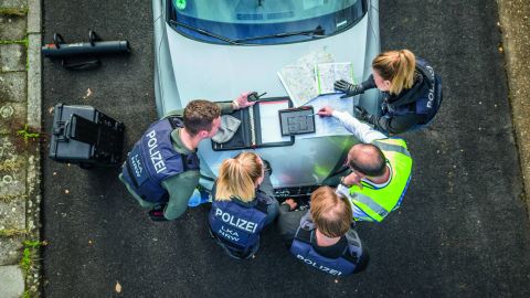 Eine Ermittlungsgruppe vom LKA NRW bestehen aus zwei Polizistinnen und drei Polizisten steht vor einem silbernen Auto. Sie haben Geräte und Landkarten auf der Motorhaube ausgebreitet und schauen darauf. Links neben dem Auto stehen weitere Gerätschaften. Das Bild wurde von oben aufgenommen.