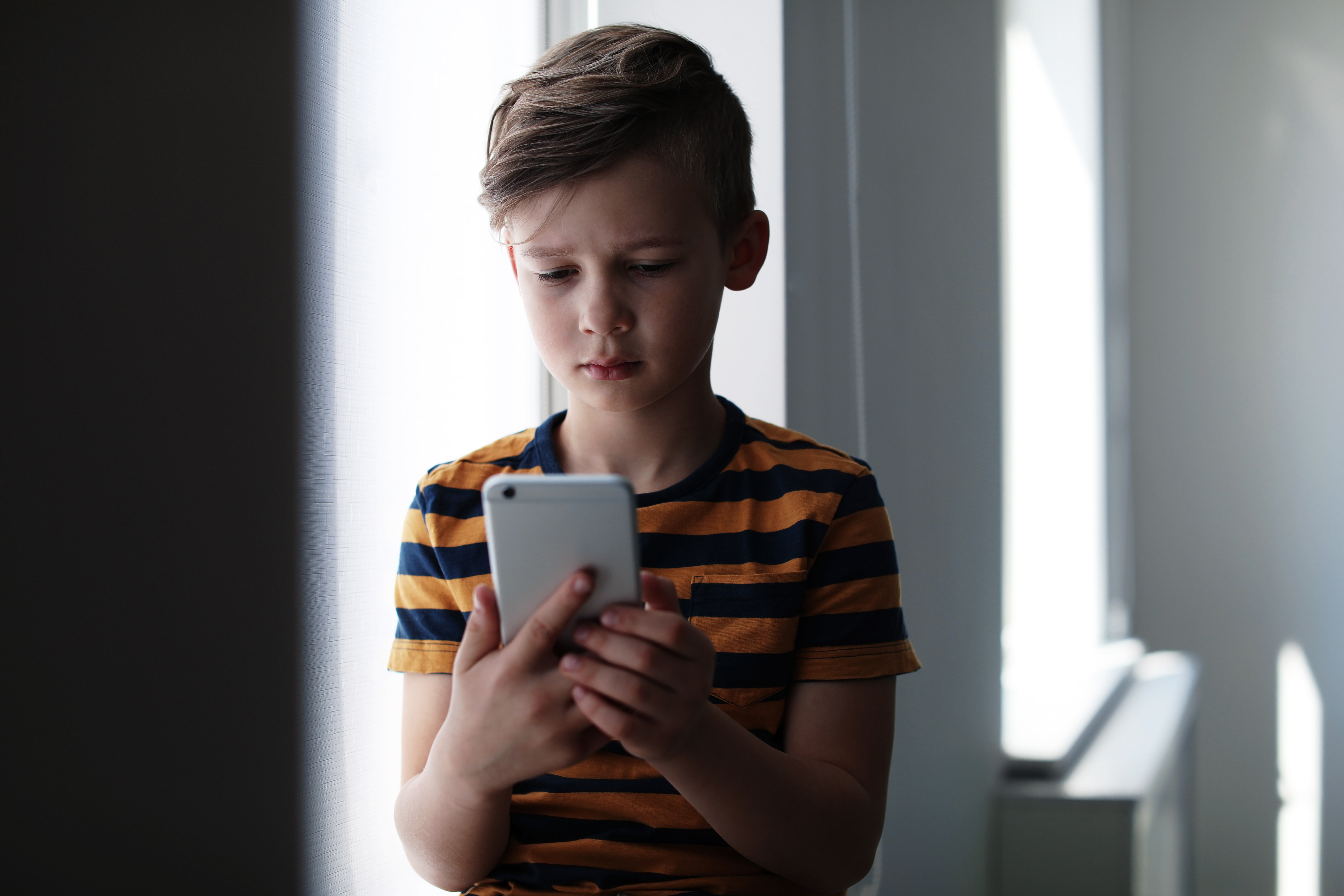 Trauriger Junge mit blau orange gestreiftem T-Shirt und weißem Smartphone in der Hand