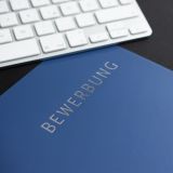 Eine blaue Bewerbungsmappe liegt auf einem Tisch mit Tastatur und Computermaus