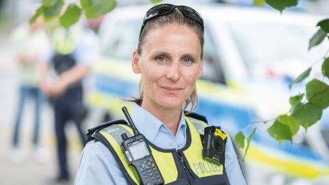 Portrait of policewoman Vanessa Göbel in uniform with blue protective vest, radio, white cap under her arm and sunglasses on her head in front of a blurred background; in the background a police car and two people