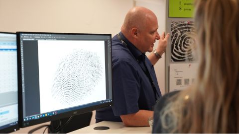 Ein Experte der Daktyloskopie ist im Hintergrund. Im Vordergrund ist ein Bildschirm mit einem Fingerabdruck. Neben ihm steht eine Stellwand mit Bildern von Fingerabdrücken. Er erläutert anhand der Bilder, wie sich der Untersuchungsbereich Daktyloskopie in den vergangenen Jahren weiterentwickelt hat.